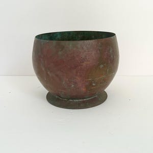 Könnte beinhalten: Eine kleine, abgerundete Kupferschale mit einer verwitterten, oxidierten Patina in rötlich-braunen und blaugrünen Farbtönen. Die Schale hat einen glatten, geschwungenen Körper, der sich zu einem runden Boden hin verjüngt und eine gealterte Metalloberfläche.