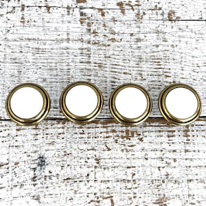 May include: Four white ceramic drawer knobs with gold metal trim, arranged in a row on a whitewashed wood surface.