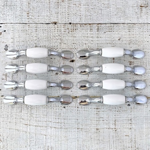 Könnte beinhalten: Sechs weiße Keramik-Schrankknöpfe mit verchromten Akzenten. Die Knöpfe sind in drei Reihen zu zweien auf einer weißen Holzoberfläche angeordnet.