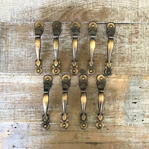 May include: Ten antique brass cabinet door pulls with a decorative design. The pulls have a round knob at the top and a pointed end at the bottom.