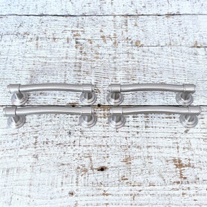 May include: Six silver-toned cabinet pulls with a curved design. The pulls are arranged in two rows of three on a white wooden surface.