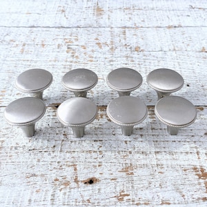 May include: Eight silver cabinet knobs with a beaded edge and a slightly pointed base. The knobs are arranged in two rows of four on a white wooden surface.