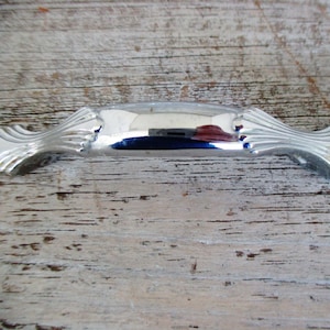 May include: A silver-toned cabinet pull with a decorative, scalloped design. The pull is curved and has a smooth, polished finish.
