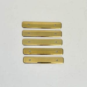 May include: Five rectangular brass plates with a polished gold-tone finish, each featuring rounded corners and two small circular holes positioned near the left and right ends for mounting purposes, arranged in a vertical stack.