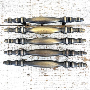 May include: Five antique bronze cabinet pulls with an ornate design. Each pull has a central handle and decorative end pieces. The pulls are arranged on a distressed white wooden surface, highlighting their vintage aesthetic. Each pull is approximately 15 cm long.