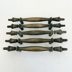 May include: A set of five horizontal drawer pulls made of aged brass metal. Each pull features a central elongated oval shape that tapers toward two decorative finials on either end, supported by two vertical mounting posts on the underside.