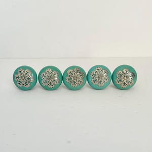 May include: Five turquoise cabinet knobs with silver filigree flower designs. These decorative knobs are round and feature a bright, solid turquoise color with a detailed silver floral overlay. Perfect for adding a touch of elegance to furniture.