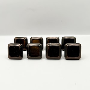 May include: Eight square cabinet knobs with a dark bronze finish are arranged in a row. The knobs have a slightly rounded shape and a smooth, reflective surface. The hardware is suitable for furniture or cabinetry.