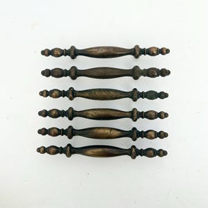 May include: A set of six matching drawer pulls arranged in a vertical stack. Each pull features a dark, aged bronze finish with a central elongated oval shape and decorative spindle-like details on both ends, creating a uniform appearance.