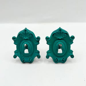 May include: Two teal-colored, ornate keyhole covers. Each cover has a decorative, scrolled design around an oval frame with a keyhole. The covers are symmetrical and appear to be made of metal.
