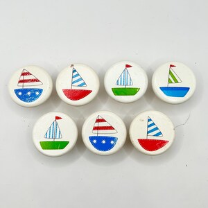 May include: A collection of eight white, round wooden buttons, each featuring a hand-painted sailboat design. The sailboats have colorful sails and hulls in red, blue, green, and blue, with small red flags. The buttons are arranged on a white surface.