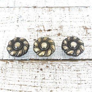 Könnte beinhalten: Drei antike goldfarbene Schrankknöpfe mit einem dekorativen floralen Design. Die Knöpfe haben eine erhabene Mitte und ein gedrehtes seilartiges Muster um die Ränder.