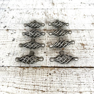 May include: Eight silver metal cabinet knobs with a twisted wire design. The knobs are arranged in two rows of four on a white wooden surface.