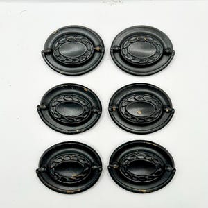 May include: Six antique black oval drawer pulls with a decorative leaf design in the center. The pulls have a dark, aged finish and are arranged in a grid pattern against a white background. Each pull has a small handle on either side.