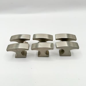 May include: Set of six silver-toned cabinet knobs with a unique T-shaped design. Each knob has a smooth, slightly curved top and a flat base.
