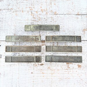 Puede incluir: Un conjunto de seis placas de metal rectangulares con acabado cepillado, dispuestas en forma de pirámide sobre una superficie de madera blanca.