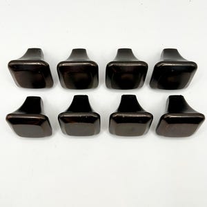 May include: Eight dark brown, geometric cabinet knobs arranged in two rows of four. Each knob has a rectangular top and a trapezoidal base. The knobs are made of a glossy material and are set against a white background.