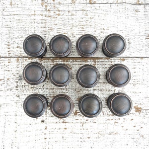 May include: Twelve round, bronze-colored cabinet knobs with a raised edge. The knobs are arranged in three rows of four on a white wooden surface.
