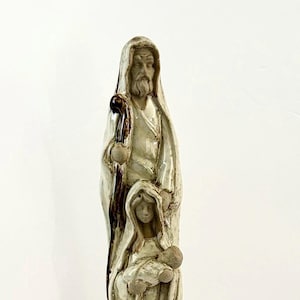 May include: A ceramic sculpture depicting a religious scene. The sculpture features three figures: a man with a beard holding a staff, a woman, and a baby. The figures are dressed in robes and have a neutral color palette.