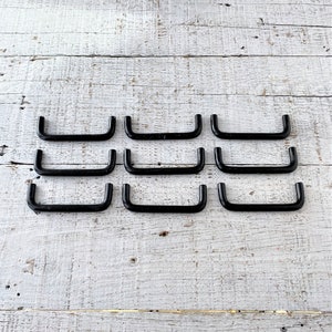 May include: Ten black metal drawer pulls in a U-shape. The pulls are arranged in three rows on a white wooden surface.
