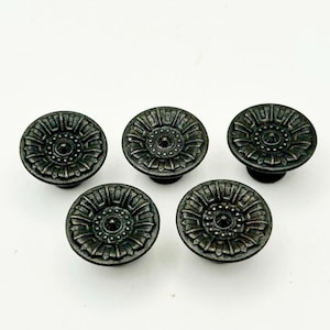 May include: Five antique-style cabinet knobs with a dark, metallic finish. Each knob features a detailed floral design with a central circular element. The knobs are arranged on a white background.