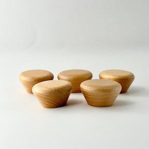 Puede incluir: Cinco pomos de madera de color marrón claro. Los pomos son redondos y tienen una parte superior ligeramente abovedada. Están dispuestos en semicírculo sobre una superficie blanca.