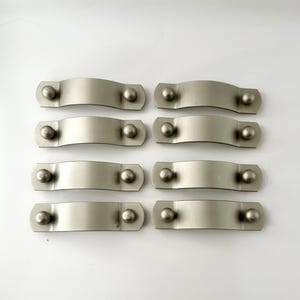 以下が含まれることがあります： 丸みを帯びたエッジとわずかに湾曲した形状の銀色のキャビネットプルが8つ。各プルには、両端に丸いノブが2つずつあります。