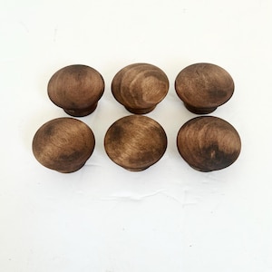 May include: A set of six round wooden knobs with a dark brown stained finish. Each knob features a smooth, domed top surface and a visible wood grain pattern, arranged in two rows of three on a plain white surface.