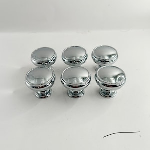 May include: Six shiny, chrome-finished cabinet knobs. Each knob has a rounded top and a decorative base. The knobs are arranged in two rows of three, reflecting light against a white background. These knobs are suitable for kitchen or bathroom cabinets.