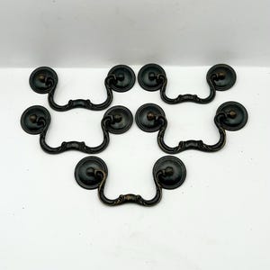 May include: Six antique bronze-toned drawer pulls with a decorative design. Each pull features a circular backplate and a curved handle. The hardware is arranged on a white background, showcasing the vintage style.