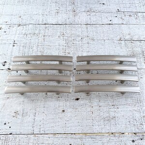 Könnte beinhalten: Acht gebürstete Nickel-Schrankgriffe mit einer geschwungenen Form und einem rechteckigen Design. Die Griffe sind in zwei Reihen zu vier auf einer weißen Holzoberfläche angeordnet.