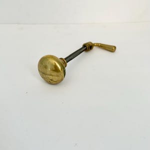 May include: A vintage brass door handle with a round knob and a lever. The handle has a dark gray shaft connecting the knob and lever. The brass has a slightly aged appearance, suggesting it is an antique.