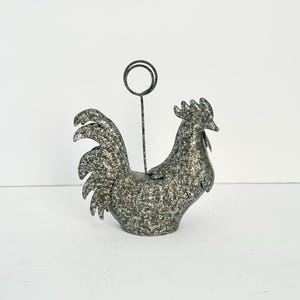 May include: A metal rooster-shaped memo holder with a speckled gray finish. The rooster has a circular wire holder extending from its back. The decorative item is designed to hold notes or photos.