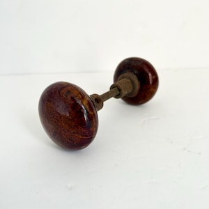 May include: A pair of antique door knobs. The knobs are a deep brown with a swirling wood grain pattern. The connecting rod and hardware are a tarnished brass color. The knobs are oval-shaped and have a glossy finish.