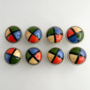 May include: Eight colorful cabinet knobs with a geometric design. Each knob features a circular, multi-colored face with a silver-toned metal rim. The colors include red, blue, green, and cream, separated by black lines. These knobs are suitable for furniture or decor.