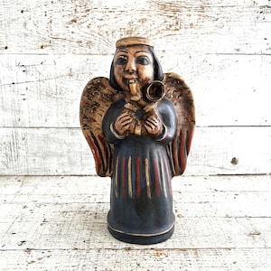 Könnte beinhalten: Geschnitzte Holzengel-Figur mit dunkelblauem Gewand und goldenen Akzenten. Der Engel hat Flügel, einen Heiligenschein und hält eine Trompete. Rote und goldene Streifen zieren das Gewand. Die Figur steht vor einem weißen Holzhintergrund.