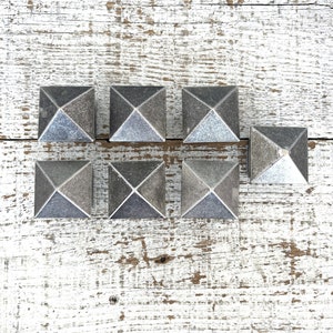 May include: Seven gray pyramid-shaped decorative knobs with a silver metallic finish. The knobs are arranged in a row on a white wooden surface.