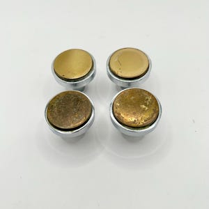 May include: Four cabinet knobs with a brass-colored top and a silver-colored base. The knobs are round and have a slightly domed shape. The brass tops show signs of wear, adding a vintage aesthetic. The knobs are arranged in a square formation.