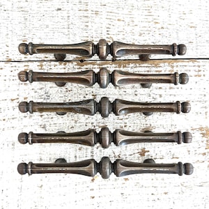 Puede incluir: Cinco tiradores de puerta de armario de bronce antiguo con diseños ornamentados. Los tiradores están dispuestos en una fila sobre un fondo de madera blanqueada.