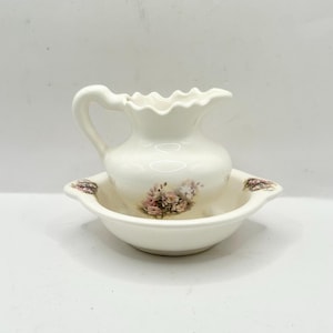 May include: A white ceramic jug and bowl set. The jug has a frilled rim and a handle. The bowl has a matching floral design with pink and purple flowers. The set is likely for decorative use.