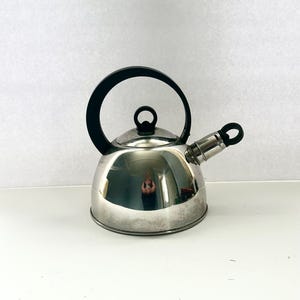 May include: A stainless steel tea kettle with a shiny, reflective surface. It has a black handle and a spout with a black tip. The kettle sits on a white surface.