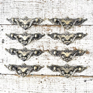 May include: Eight ornate brass drawer pulls with a decorative design. The pulls have a curved shape and are finished in a polished brass color.