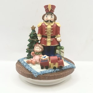 Può includere: Statuetta natalizia decorativa raffigurante un soldatino con cappotto rosso e cappello alto. La statuetta include un piccolo albero di Natale, una bambola, un trenino giocattolo e un regalo confezionato. La base è marrone chiaro con bordi blu.