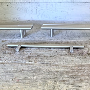 Może przedstawiać: Trzy srebrne uchwyty do szafek z prostokątnym wzorem. Uchwyty znajdują się na jasnym drewnianym blacie.