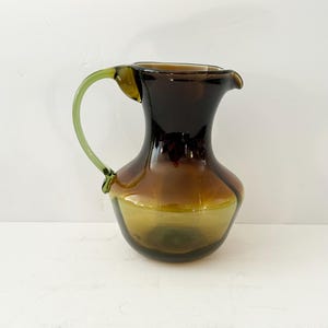 Può includere: Una brocca decorativa in vetro con una parte superiore marrone scuro che sfuma in un marrone più chiaro e poi in una base verde. Il manico è di colore verde chiaro. La brocca ha una forma e un design unici.