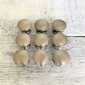 May include: Ten silver cabinet knobs with a round, slightly raised design. The knobs are arranged in three rows on a white wooden surface.