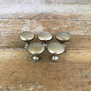 Puede incluir: Seis pomos de armario plateados con forma redonda y un pequeño tallo. Los pomos están dispuestos en semicírculo sobre una superficie de madera.