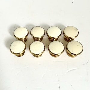 May include: A set of eight round cabinet knobs arranged in two rows of four. Each knob features a smooth, off-white circular top surface encased in a polished gold-tone metal rim and base, creating a uniform appearance for furniture hardware.