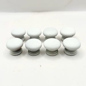 May include: Eight white, round cabinet knobs with a speckled texture. The knobs have a classic mushroom shape, with a rounded top and a cylindrical base. They are arranged in two rows of four against a white background.