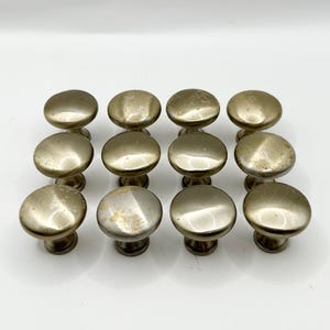 May include: A set of 12 silver-toned cabinet knobs with a round, slightly domed shape. The knobs have a brushed finish and a subtle aged look.